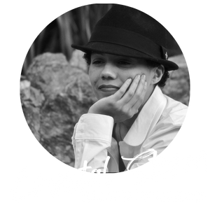 Voice Actor and Voice Director Crystal Storm. Black and White photo. Crystal is wearing a fedora, with her hand against her face, staring off into the distance.
